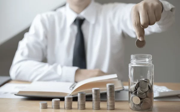 Person in weißem Hemd legt Münze in ein Glas voller Geld neben gestapelten Münzsäulen – Symbol für Geld sparen im Alltag.